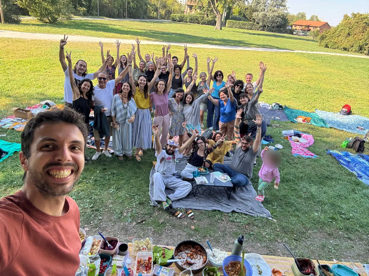 Un ragazzo sorride in un selfie; dietro di lui il gruppo, nel parco, alza le braccia